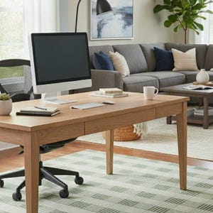 May include: A light wood desk with a computer monitor, keyboard, and lamp sits on a green and white plaid rug. A grey sectional sofa with cushions and a painting are in the background. The room has a modern, minimalist design.