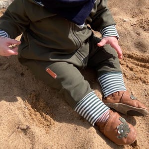 Könnte beinhalten: Olivgrüner Baby-Overall mit passender Jacke, gestreiften Bündchen und einem kleinen Aufnäher. Dazu braune Lederbabyschuhe mit grünem Applikation.