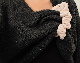 Könnte beinhalten: Ein hellrosa Stoff-Haargummi mit Waffelstruktur und einem Muster aus kleinen braunen Herzen, das auf einem dunkel anthrazitfarbenen Strickpullover mit V-Ausschnitt und geripptem Kragendetail liegt.