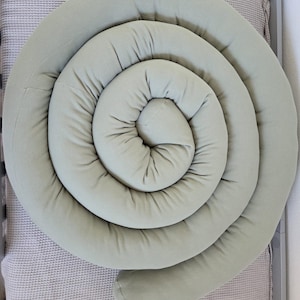 Könnte beinhalten: Ein spiralförmiger, salbeigrüner Bettumrandung. Der Bumper besteht aus einem weichen, strukturierten Stoff und ist so konzipiert, dass er sich um die Innenseite eines Kinderbettes wickelt. Der Bumper ist einfarbig und hat eine abgerundete Form.