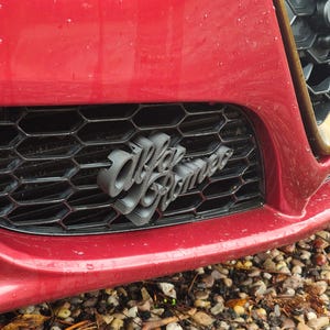 Op de afbeelding: Close-up van de voorkant van een rode auto, met een zwarte honingraatgrille en het grijze "Alfa Romeo" embleem. De carrosserie van de auto heeft een glanzende afwerking, het embleem is duidelijk zichtbaar.