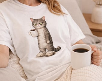 May include: White t-shirt featuring a grumpy cartoon cat holding a coffee cup. The cat is gray and white with yellow eyes. The shirt is a casual style, perfect for cat lovers.
