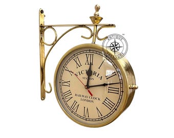 Könnte beinhalten: Eine doppelseitige Eisenbahn-Wanduhr aus Messing mit dem Text "VICTORIA STATION" und "RAILWAY CLOCK LONDON" auf dem cremefarbenen Zifferblatt. Die Uhr hat schwarze römische Ziffern und Zeiger sowie einen roten Sekundenzeiger. Die Uhr ist an einer Messinghalterung befestigt.