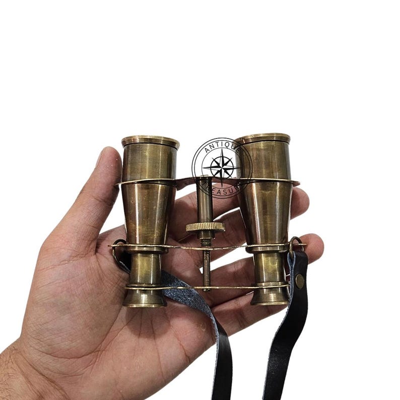 K&ouml;nnte beinhalten: Antikes Messingfernglas mit einem schwarzen Lederriemen. Das Fernglas hat ein klassisches Design mit einstellbarem Fokus und eine Vintage-&Auml;sthetik. Das Bild zeigt das Fernglas in einer Hand vor wei&szlig;em Hintergrund.