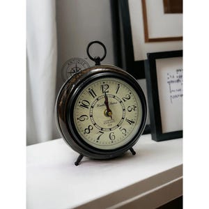 May include: Antique-style round clock with a black metal frame and a cream-colored face. The clock features black Roman numerals, a small handle on top, and a base with three small feet. The clock face reads "Hunting Station, Cambridge England 1924."