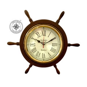 Puede incluir: Un reloj de timón de barco de madera marrón con un bisel dorado y una esfera crema. La esfera del reloj presenta números romanos negros y las palabras "Das Santos London". El reloj tiene seis radios y un diseño vintage. El diámetro es de unos 30 cm.