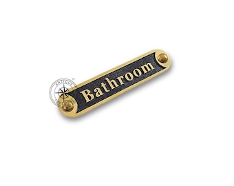 May include: A rectangular bathroom sign with the word "Bathroom" in gold lettering on a black background. The sign has gold-colored rounded ends with a screw head on each end. The sign is a decorative item.