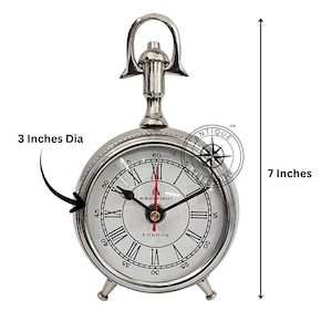 May include: A silver-coloured, round clock with a handle on top and three small feet. The clock face has Roman numerals and the word "LONDON". The clock is 17.78 cm tall and 7.62 cm in diameter.