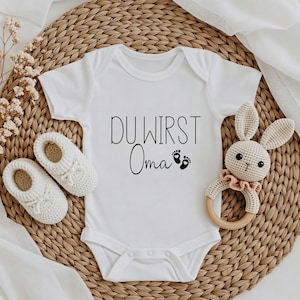 May include: White baby onesie with the text "DU WIRST Oma" in black script, along with two small footprints. The onesie is surrounded by white crochet baby shoes, a crochet bunny rattle, and a woven mat.