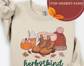 Könnte beinhalten: Beiges Sweatshirt mit einem herbstlichen Motiv. Das Design zeigt einen Kürbis, Stiefel, eine Mütze und ein Kaffeegetränk. Der Text "herbstkind EST. 2005" ist darunter gedruckt. Ein kreisförmiges Grafik mit deutschem Text befindet sich oben rechts.
