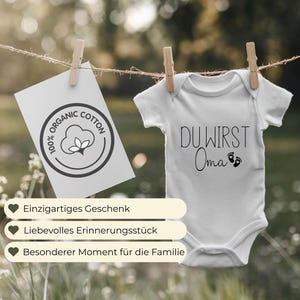 May include: A white baby onesie with the text "DU WIRST Oma" and tiny footprints. A card with "ORGANIC COTTON" text and a cotton flower logo hangs nearby. The image also shows text in German: "Einzigartiges Geschenk", "Liebevolles Erinnerungsst&uuml;ck", and "Besonderer Moment f&uuml;r die Familie".