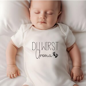May include: A sleeping baby wearing a white onesie with the text "DU WIRST Uroma" in black script, along with two small foot prints. The baby is lying on a white pillow and sheet.