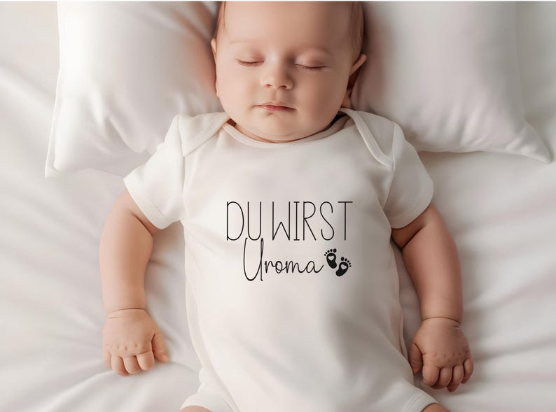 May include: A sleeping baby wearing a white onesie with the text "DU WIRST Uroma" in black script, along with two small foot prints. The baby is lying on a white pillow and sheet.