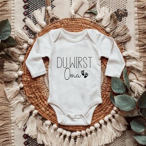 May include: White long-sleeved baby onesie with the German text "DU WIRST Oma" and two small black footprints. The onesie is laid on a woven basket and a patterned rug.
