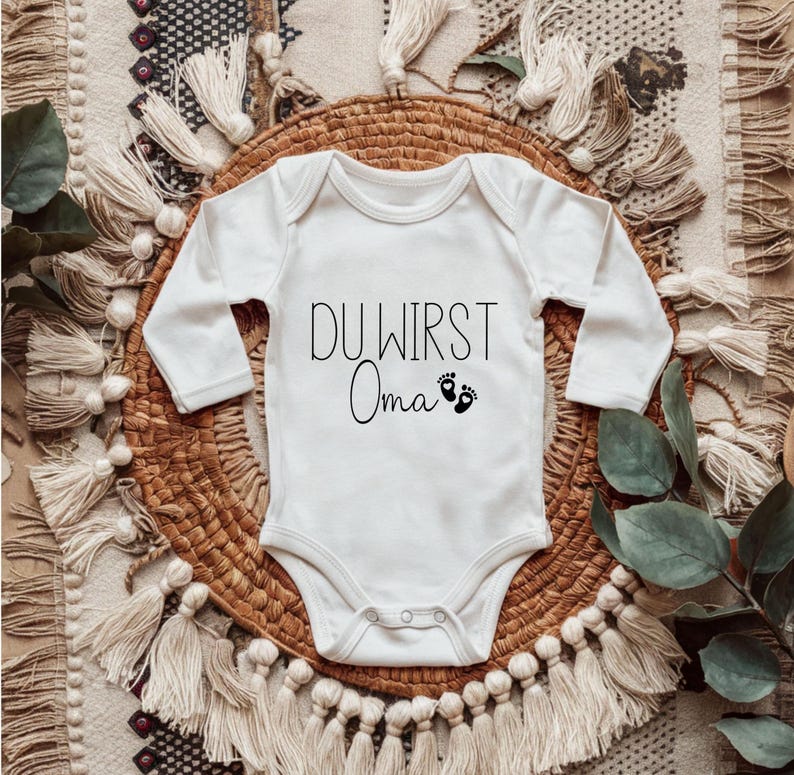 May include: White long-sleeved baby onesie with the German text "DU WIRST Oma" and two small black footprints. The onesie is laid on a woven basket and a patterned rug.