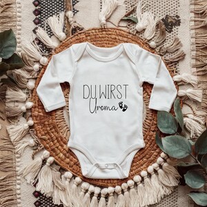 May include: White baby onesie with long sleeves. The text "DU WIRST Uroma" is printed in black script, along with two small footprints. The onesie is placed on a woven basket with a decorative textile background.