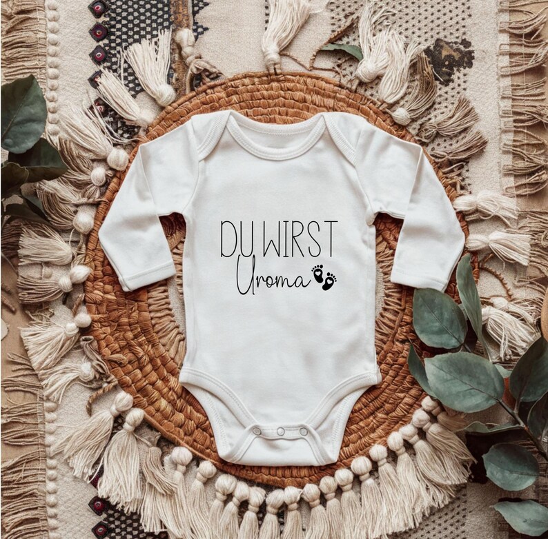 May include: White baby onesie with long sleeves. The text "DU WIRST Uroma" is printed in black script, along with two small footprints. The onesie is placed on a woven basket with a decorative textile background.