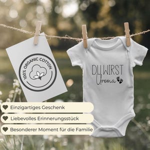 May include: A white baby onesie with the text "DU WIRST Uroma" in black script, hanging from a clothesline. A card with "100% ORGANIC COTTON" is clipped to the line. Wooden clothespins hold the items. The background is blurred.