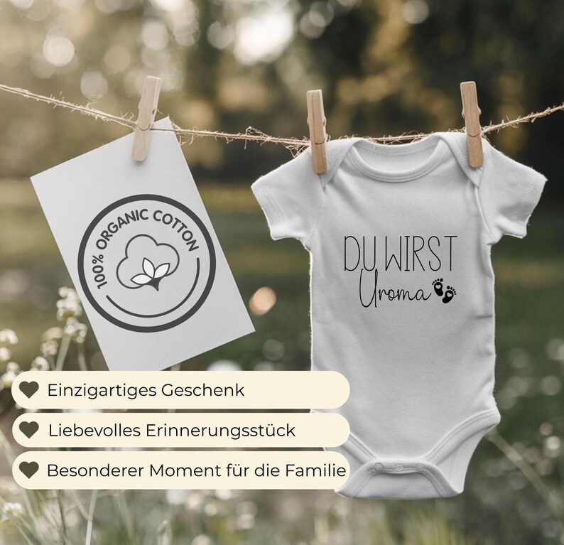 May include: A white baby onesie with the text "DU WIRST Uroma" in black script, hanging from a clothesline. A card with "100% ORGANIC COTTON" is clipped to the line. Wooden clothespins hold the items. The background is blurred.
