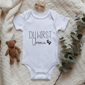 May include: White baby onesie with the text "DU WIRST Uroma" in black script, along with two small black footprints. A small teddy bear and a beige bow are visible in the background. The onesie is a newborn garment.