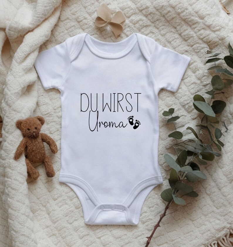 May include: White baby onesie with the text "DU WIRST Uroma" in black script, along with two small black footprints. A small teddy bear and a beige bow are visible in the background. The onesie is a newborn garment.
