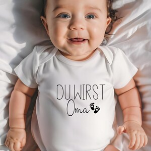 May include: A smiling baby wearing a white onesie with the German text "DU WIRST Oma" and two small foot prints. The baby is lying on a white sheet.
