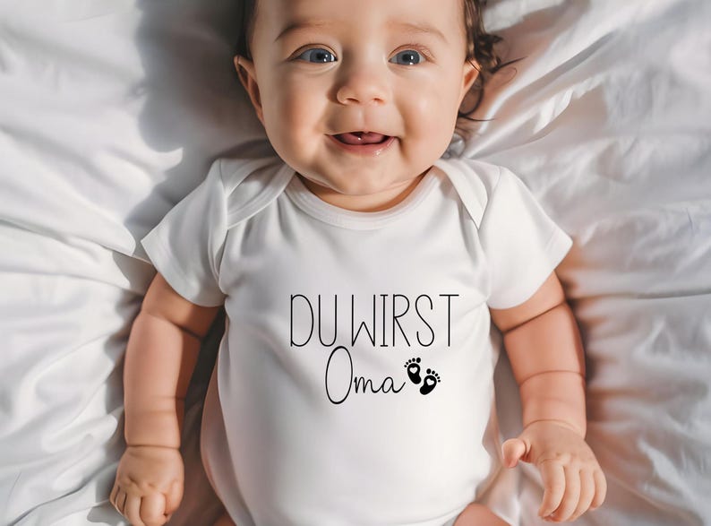 May include: A smiling baby wearing a white onesie with the German text "DU WIRST Oma" and two small foot prints. The baby is lying on a white sheet.