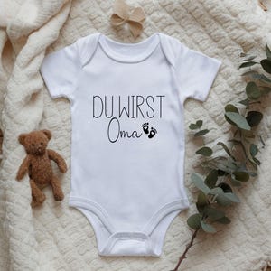 May include: White baby onesie with the text "DU WIRST Oma" in black script, along with two small black footprints. A small brown teddy bear and a beige bow are also in the image.