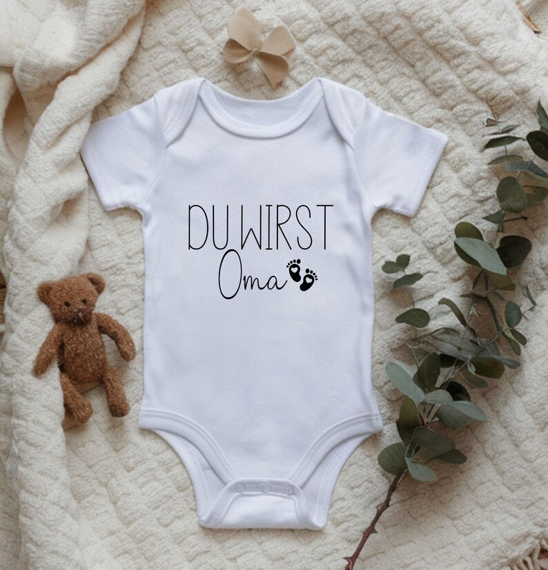 May include: White baby onesie with the text "DU WIRST Oma" in black script, along with two small black footprints. A small brown teddy bear and a beige bow are also in the image.