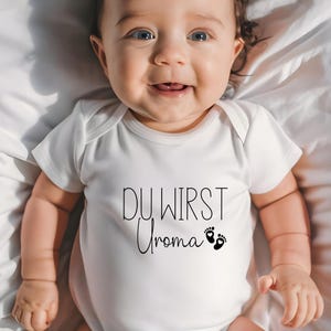 May include: A smiling baby wearing a white onesie with the German text "DU WIRST Uroma" and two black footprints. The baby is lying on a white sheet.