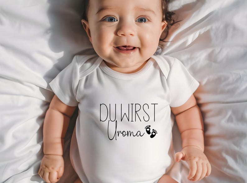May include: A smiling baby wearing a white onesie with the German text "DU WIRST Uroma" and two black footprints. The baby is lying on a white sheet.