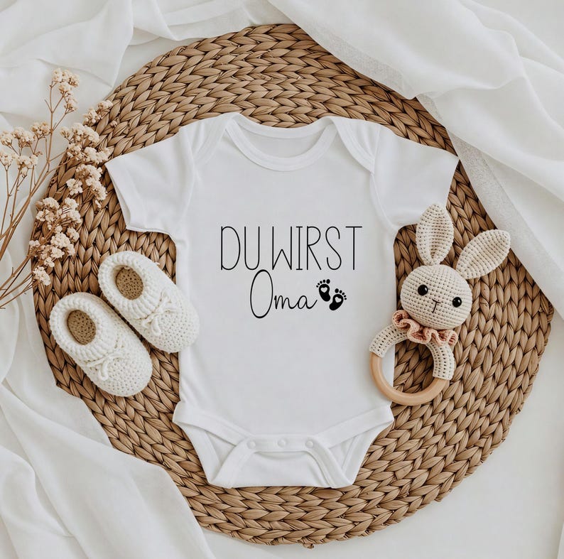 May include: White baby onesie with the text "DU WIRST Oma" in black script, along with two small footprints. The onesie is surrounded by white crochet baby shoes, a crochet bunny rattle, and a woven mat.