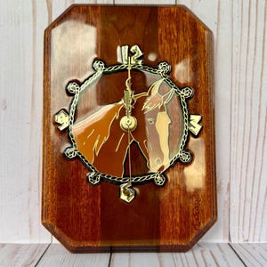 May include: A rectangular wooden wall clock with a horse head design. The clock face features a brown horse head within a gold-colored frame. The clock hands and numerals are gold. The wooden base has a glossy finish.