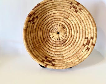 May include: A round, woven basket with a natural, light brown color. The basket features concentric circles radiating from the center, with dark brown geometric patterns around the rim. The basket is elevated on small, black feet.