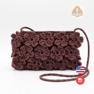 May include: A dark brown rectangular crochet bag featuring a dense pattern of three-dimensional crocheted flowers across the front surface. The bag includes a long, thin braided shoulder strap and displays the text magic crochet verse.