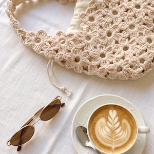 Puede incluir: Un bolso de ganchillo beige con patr&oacute;n floral y cierre de cord&oacute;n, unas gafas de sol con montura dorada y cristales marrones, y una taza de cer&aacute;mica blanca con caf&eacute; con arte latte sobre un plato a juego con una cuchara de metal plateado.