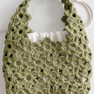 Puede incluir: Un bolso de ganchillo color verde salvia con un patr&oacute;n de motivos florales repetidos en todo el cuerpo y el asa. El bolso incluye un forro de tela blanca con cord&oacute;n que es visible a trav&eacute;s del dise&ntilde;o de ganchillo calado exterior.
