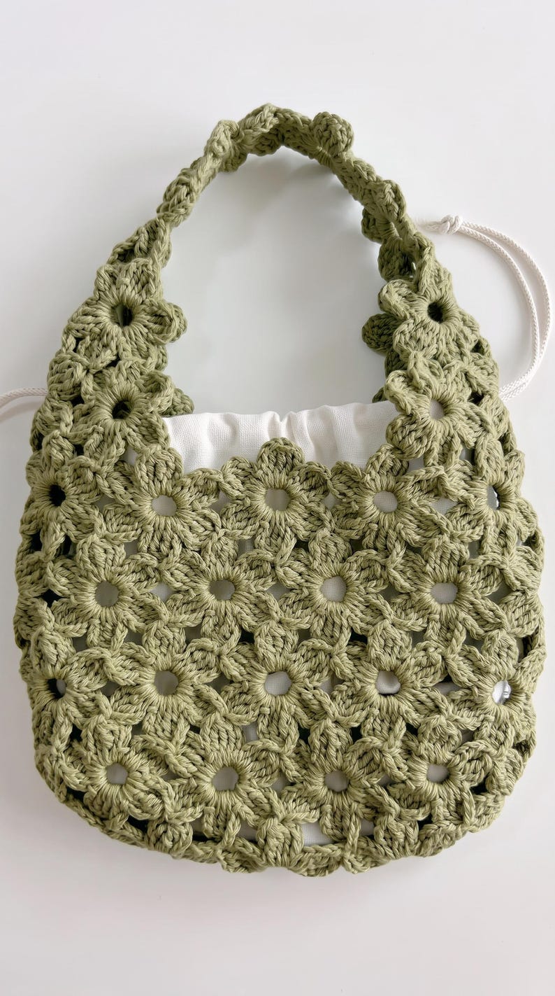 Puede incluir: Un bolso de ganchillo color verde salvia con un patr&oacute;n de motivos florales repetidos en todo el cuerpo y el asa. El bolso incluye un forro de tela blanca con cord&oacute;n que es visible a trav&eacute;s del dise&ntilde;o de ganchillo calado exterior.