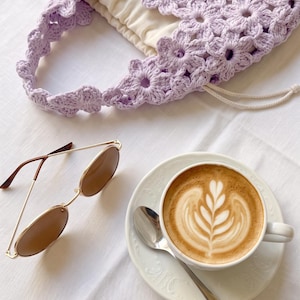 Puede incluir: Un bolso de ganchillo de color violeta claro con patr&oacute;n floral, forro de tela color crema y cierre de cord&oacute;n, gafas de sol con montura dorada y cristales marrones, y una taza de cer&aacute;mica blanca con un latte decorado con arte en espuma.