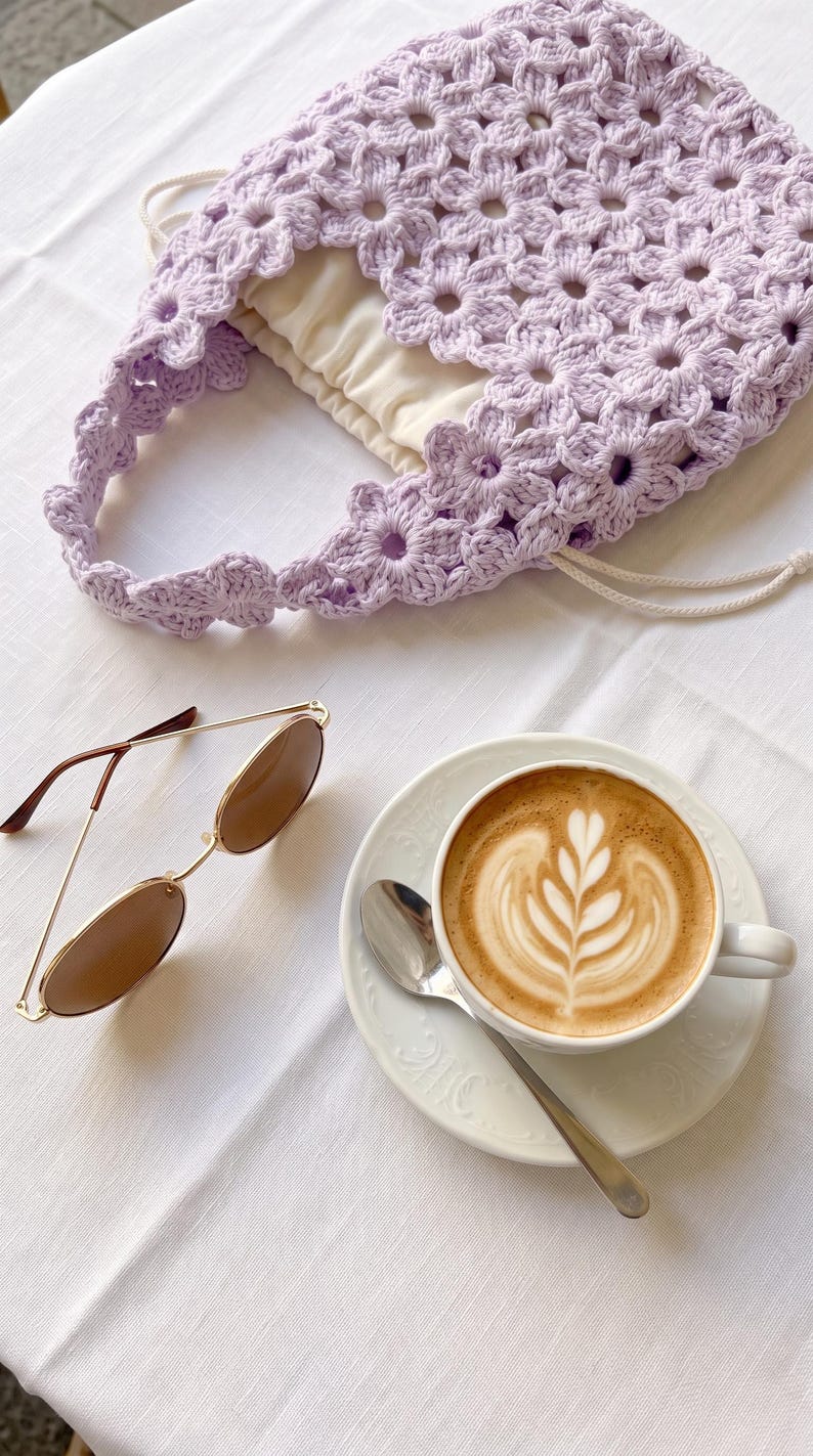 Puede incluir: Un bolso de ganchillo de color violeta claro con patr&oacute;n floral, forro de tela color crema y cierre de cord&oacute;n, gafas de sol con montura dorada y cristales marrones, y una taza de cer&aacute;mica blanca con un latte decorado con arte en espuma.