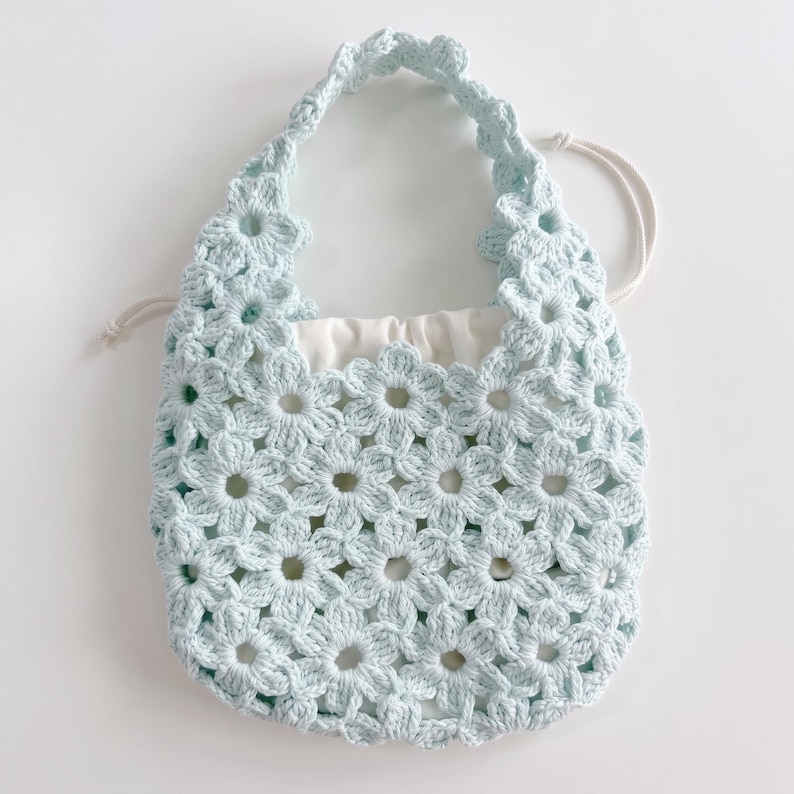 Puede incluir: Un bolso de ganchillo azul claro construido con motivos florales interconectados. El bolso cuenta con un asa integrada &uacute;nica y un forro de tela blanca con cord&oacute;n que se ajusta en la parte superior para asegurar el contenido del dise&ntilde;o calado.