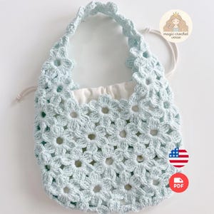 Puede incluir: Un bolso de ganchillo azul claro con un patr&oacute;n de motivos florales repetidos en todo el cuerpo y el asa. El bolso incluye un forro interior de tela blanca con cord&oacute;n y un logotipo circular en la esquina superior derecha que dice magic crochet verse.