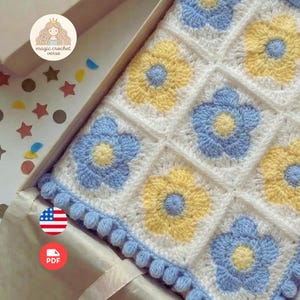 May include: A handmade crochet blanket featuring a floral pattern in white, yellow, and light blue. The blanket has a border of blue pom-poms. The blanket is inside a box. The text "magic crochet verse" is visible.