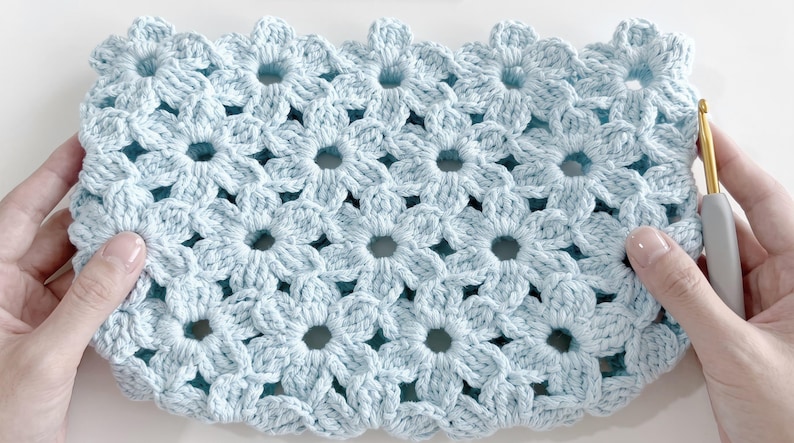 Puede incluir: Un tejido de ganchillo azul claro con un patr&oacute;n floral repetitivo hecho de motivos de flores interconectados. El textil tiene un dise&ntilde;o calado con centros circulares y bordes en forma de p&eacute;talos. Un ganchillo gris y dorado se sit&uacute;a a la derecha.