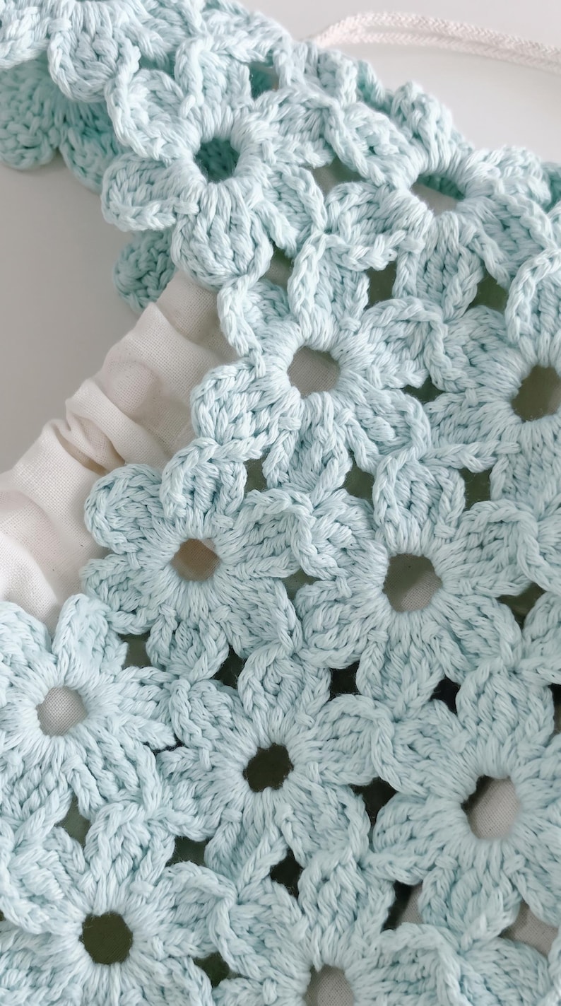 Puede incluir: Un bolso de ganchillo azul claro con un patr&oacute;n de motivos florales repetitivos. El bolso est&aacute; construido con flores de hilo interconectadas e incluye un forro de tela blanca visible a trav&eacute;s del dise&ntilde;o calado, junto con un cord&oacute;n blanco.