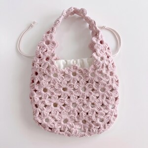 Puede incluir: Un bolso de mano de ganchillo en color rosa claro construido con un patr&oacute;n de motivos florales repetitivos. El bolso cuenta con un asa integrada &uacute;nica y un forro de tela blanca con cord&oacute;n que se ajusta en la parte superior para asegurar el contenido.