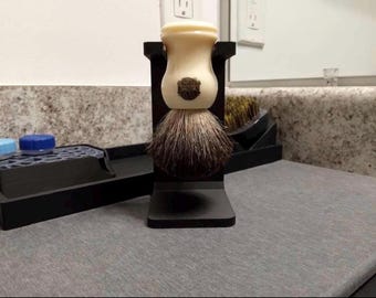 May include: A shaving brush with a cream-coloured handle and dark bristles is displayed in a black stand. The brush is the focal point, with a small emblem on the handle. Other grooming accessories are visible in the background.