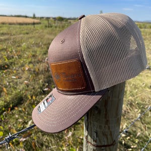 May include: A brown baseball cap with a beige mesh back and a brown leather-like patch on the front. The patch features the text BB CONCEPTS CANADA. The cap has a curved brim with visible stitching and a small rectangular tag attached to the side.
