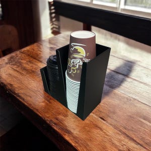 Può includere: Un organizer nero per tazze e coperchi da caffè su un tavolo di legno. Contiene pile di tazze da caffè marroni con bordo bianco con un design a tema caffè e una pila di coperchi neri. Lo sfondo è sfocato.