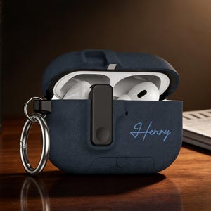 May include: Navy blue AirPods case with the name "Henry" in a script font. The case is open, revealing white AirPods. A silver keyring is attached to the case. The case is sitting on a wooden surface.